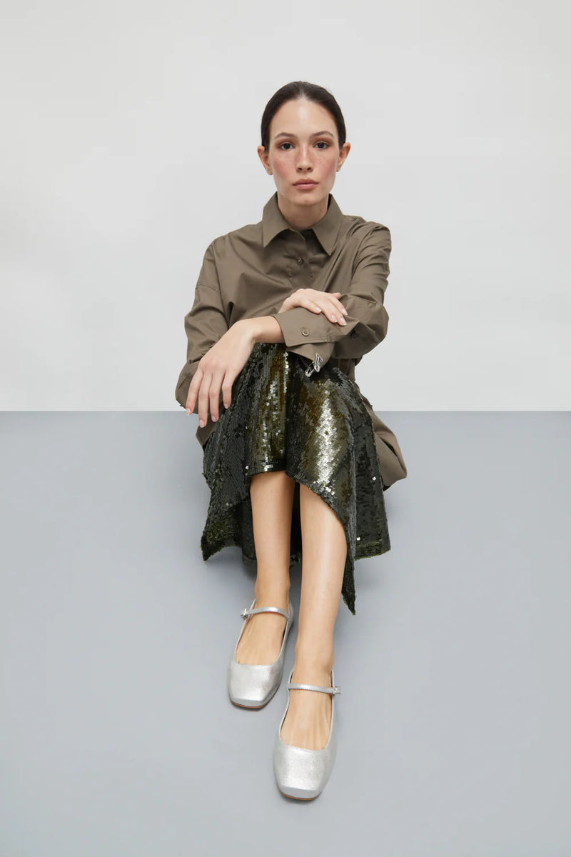 Woman sitting on a gray surface wearing a brown shirt and green sequin skirt.