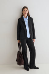 A woman stands against a plain light gray studio wall wearing a dark charcoal plaid blazer and matching wide-leg trousers over a light blue button-up shirt, holding a dark brown suede tote bag.