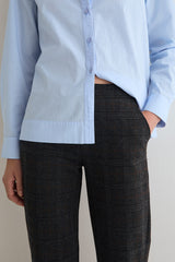 A person wearing a light blue long-sleeve button-up shirt and dark charcoal plaid trousers, shown from the chest down against a plain light beige background.
