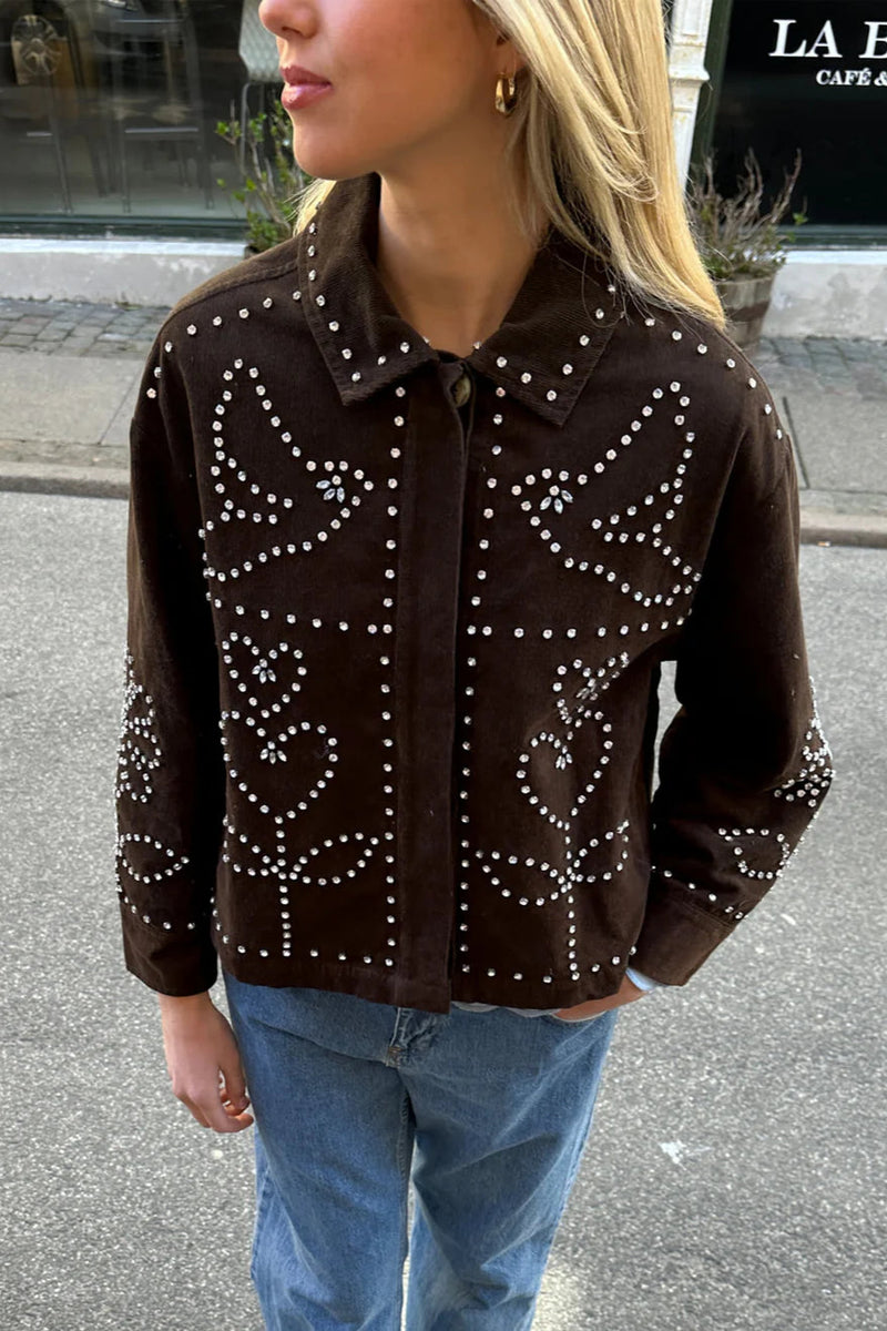Person wearing a brown embellished jacket with blue jeans on a street.