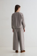 A model viewed from the back wearing a matching light gray knit lounge set consisting of a crewneck sweatshirt and wide-leg trousers, paired with brown slippers on a clean white studio background.