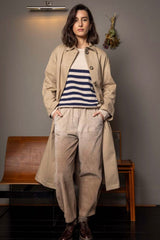 Woman wearing a beige trench coat and striped shirt in a room with wooden furniture.