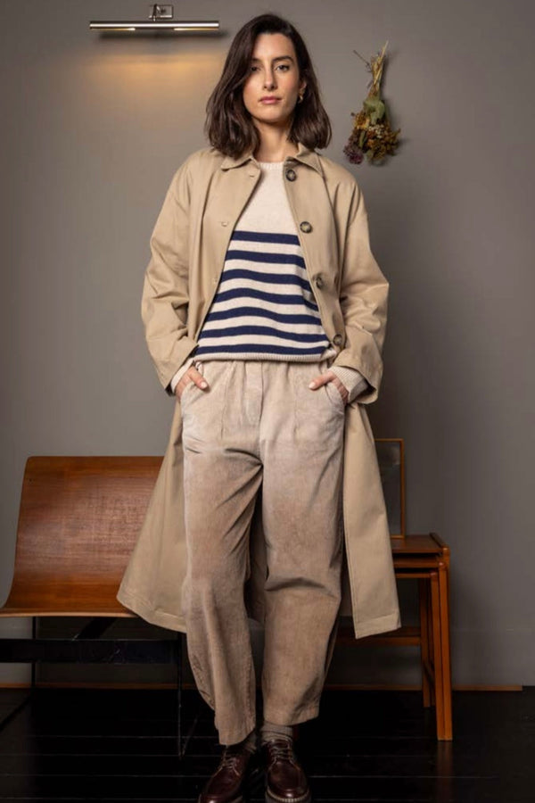 Woman wearing a beige trench coat and striped shirt in a room with wooden furniture.