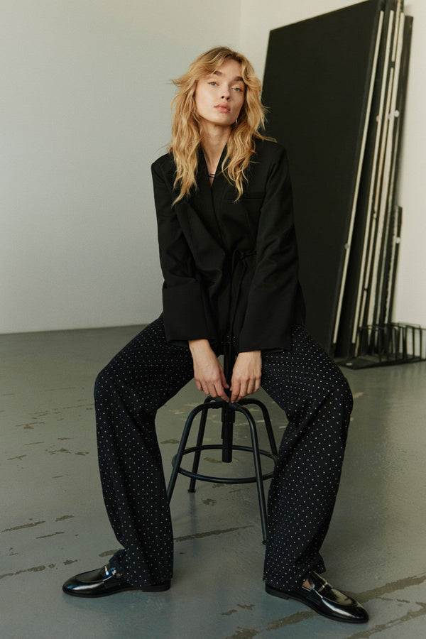 Person wearing a black blazer and polka dot pants sitting on a stool in a minimalistic setting.