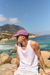 Person wearing a white tank top and purple cap sitting on rocks by a scenic coastal area with mountains and blue sky.